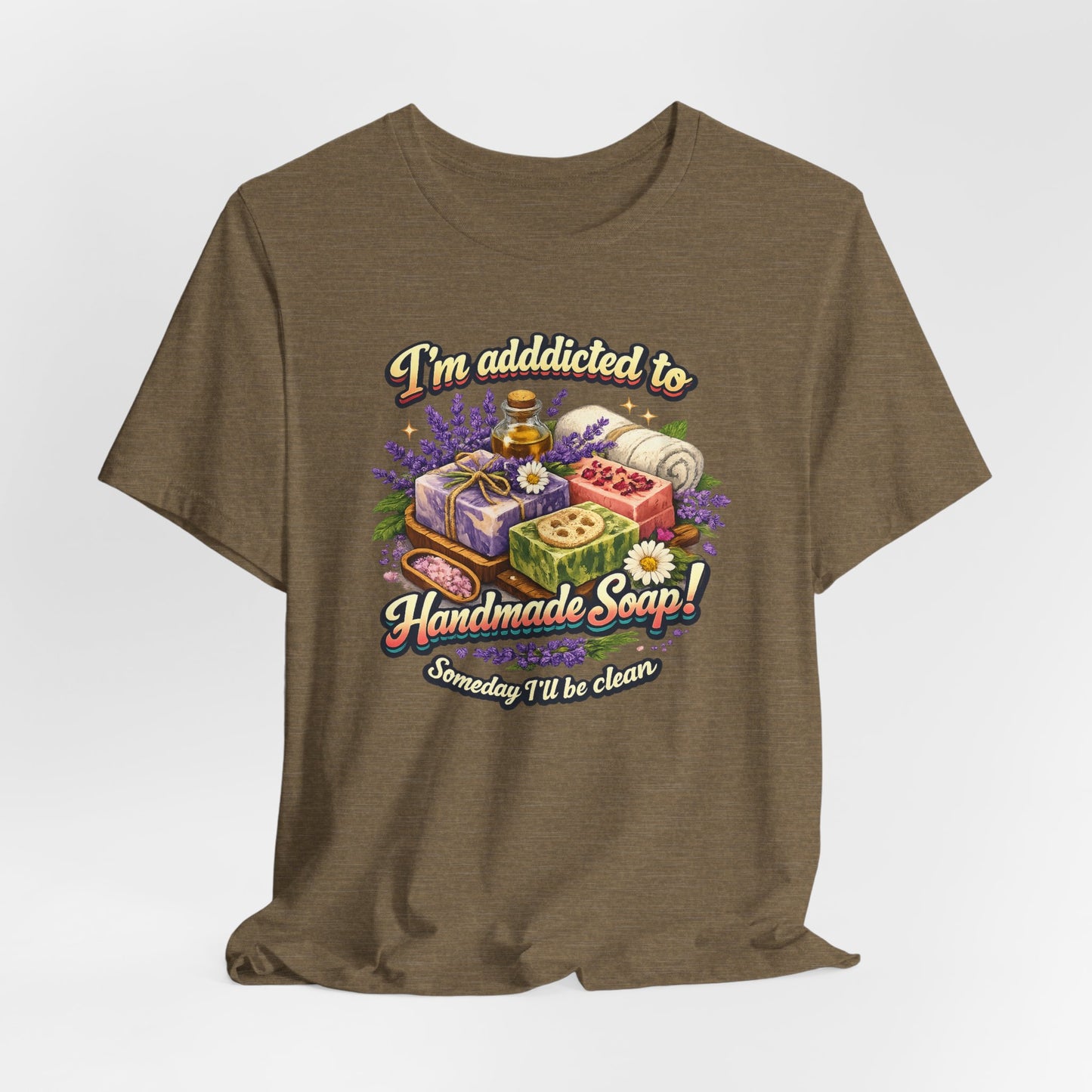 A brown t-shirt displays a colorful graphic stating "I'm addicted to Handmade Soap! Someday I'll be clean," featuring various artisan soaps, lavender, and essential oils.
