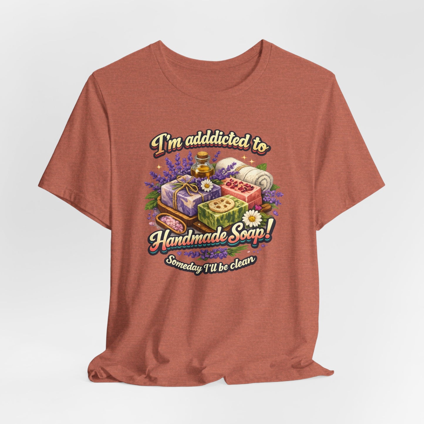 Heather coral t-shirt displays a whimsical design featuring handmade soap, lavender, and the saying "I'm addicted to Handmade Soap! Someday I'll be clean."
