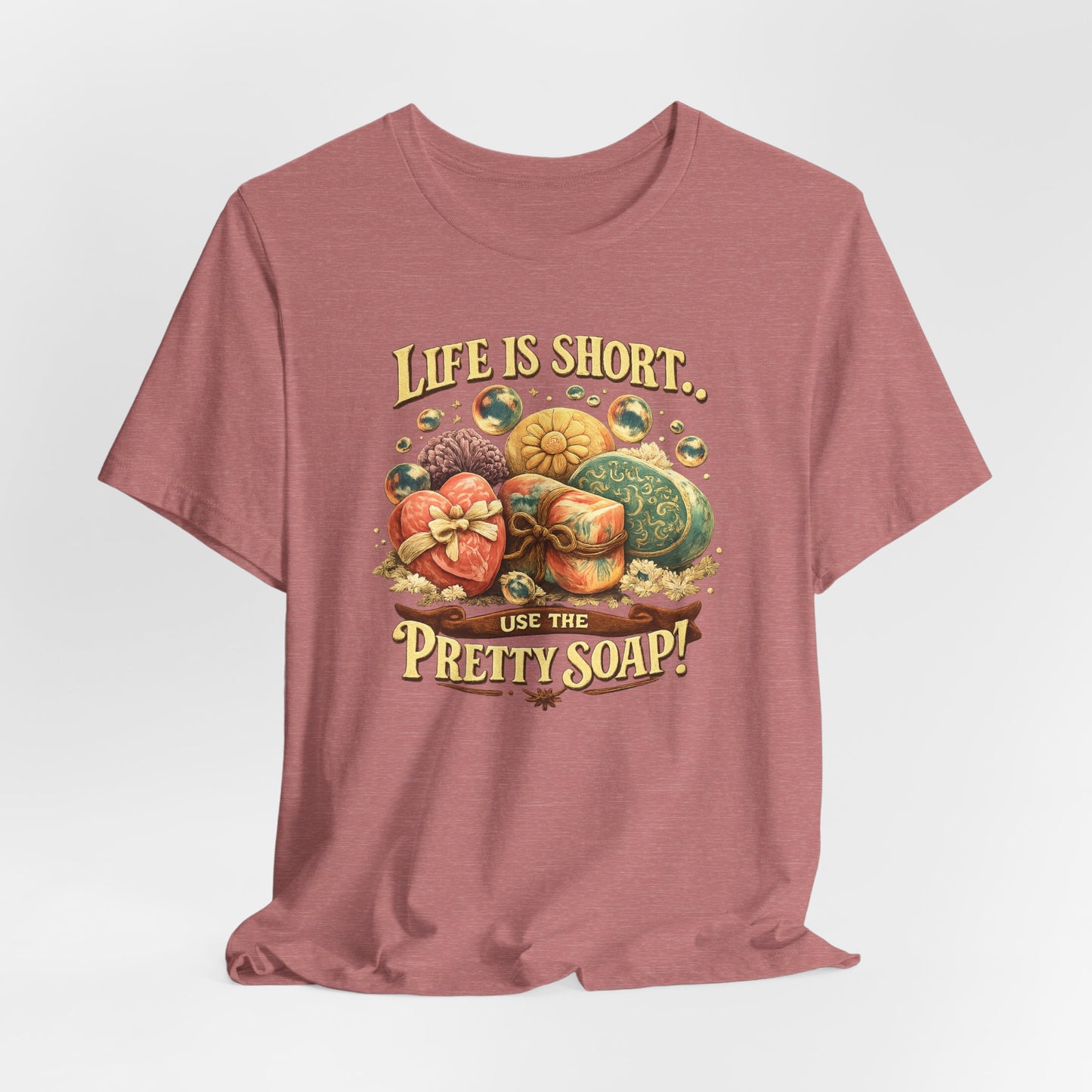 Displays a heathered rose-colored T-shirt featuring decorative soaps and the quote "Life is short, use the pretty soap!"
