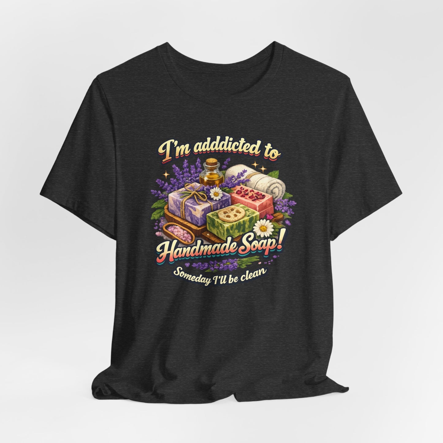 Person wears a dark gray t-shirt featuring an illustration of colorful handmade soaps, lavender, and a witty saying about soap addiction.

