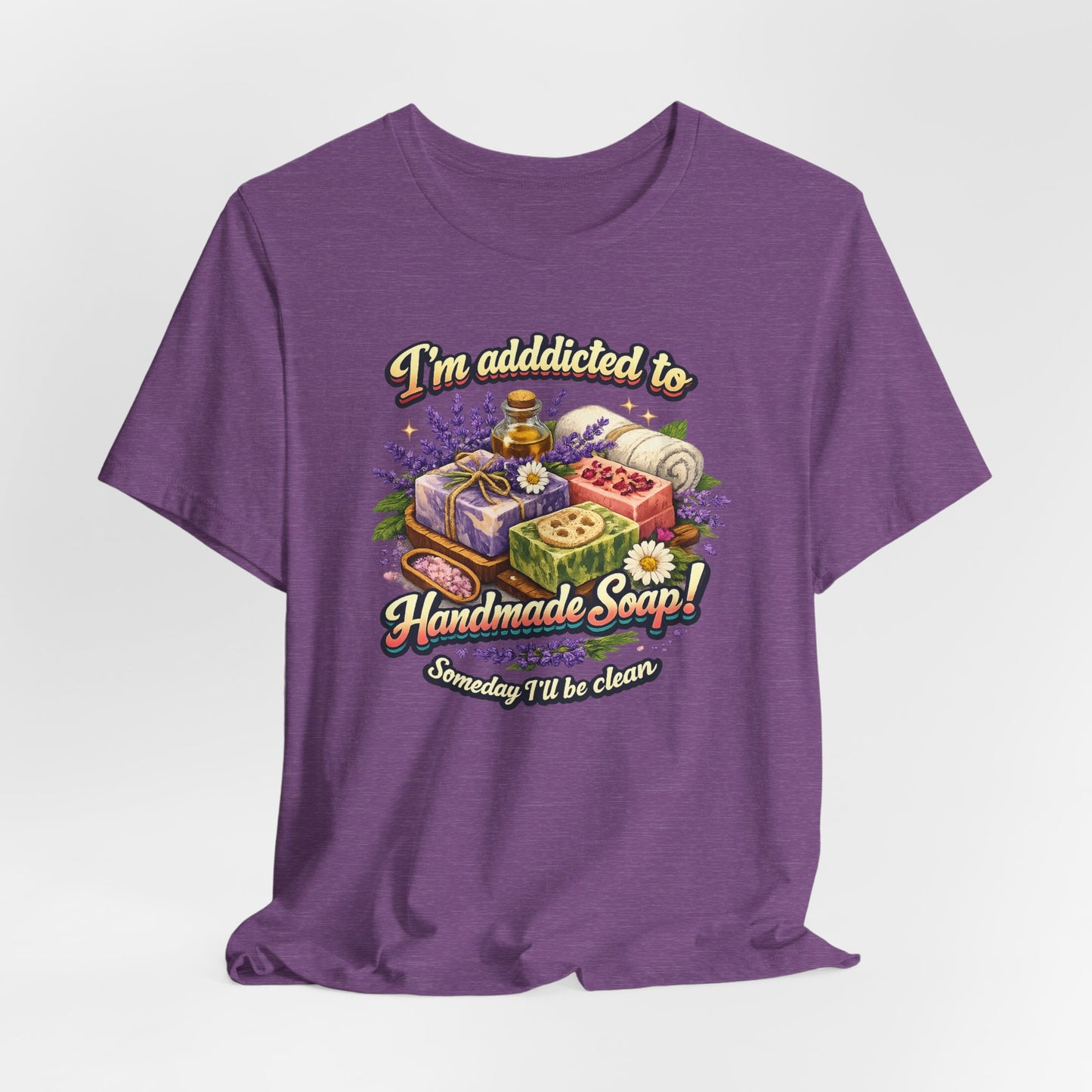 A purple t-shirt displays a colorful graphic promoting handmade soap with lavender and a humorous message about being clean someday.
