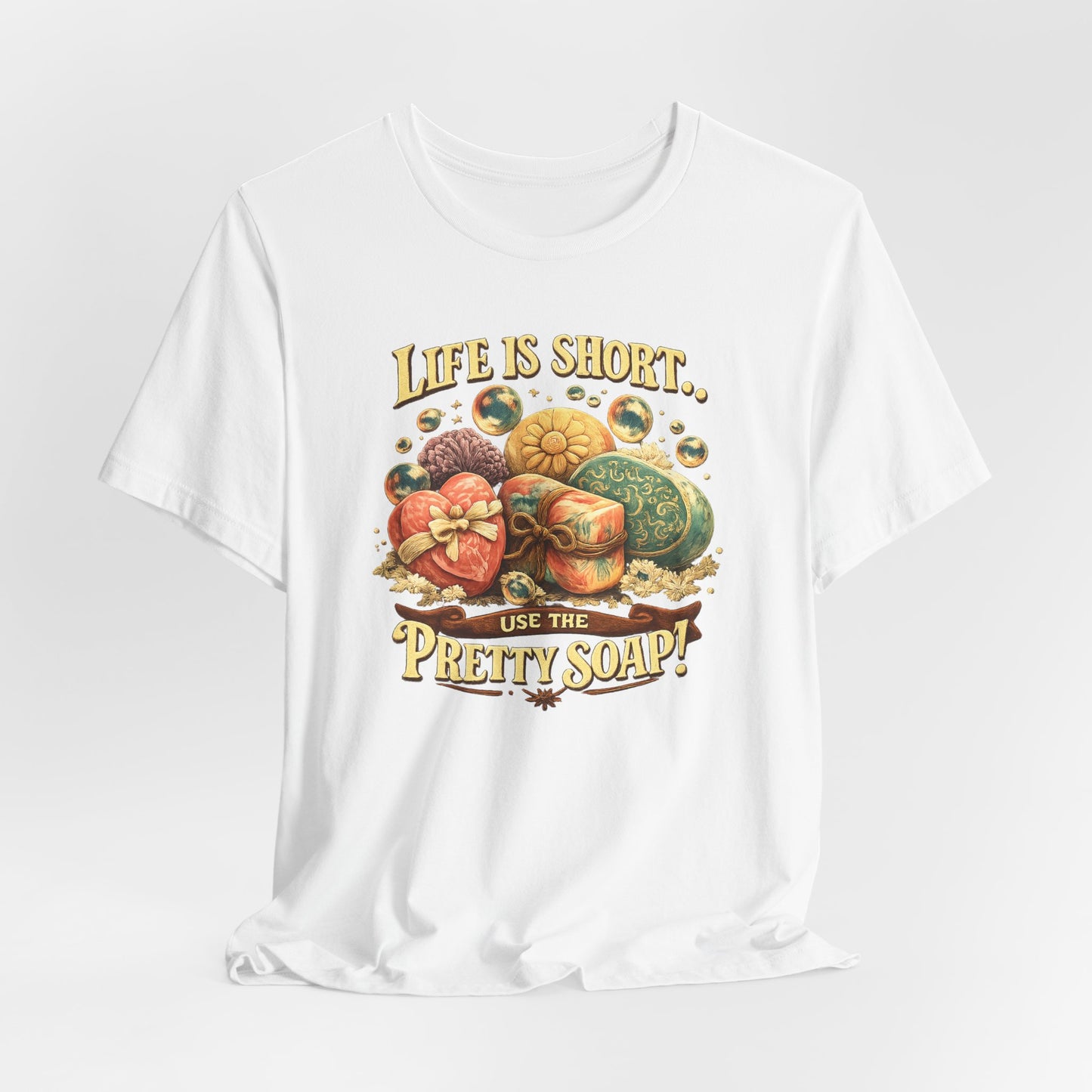 Shows a white t-shirt featuring a graphic design promoting the use of pretty soap, with a variety of decorative soaps and the message "Life is short, use the pretty soap!".
