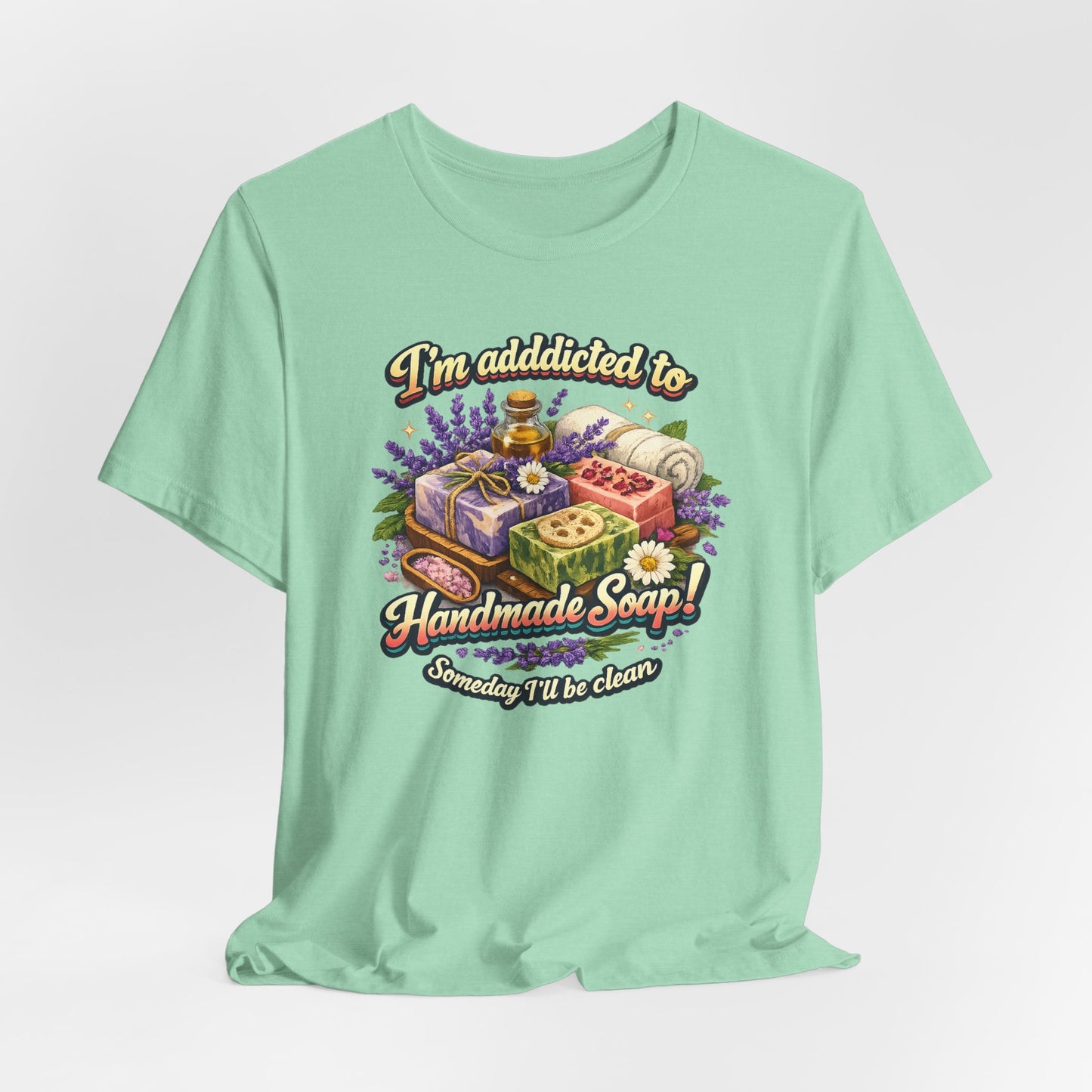 A light green t-shirt displays the whimsical text "I'm addicted to Handmade Soap! Someday I'll be clean" above an illustration of artisan soaps, essential oil, lavender, and a towel.
